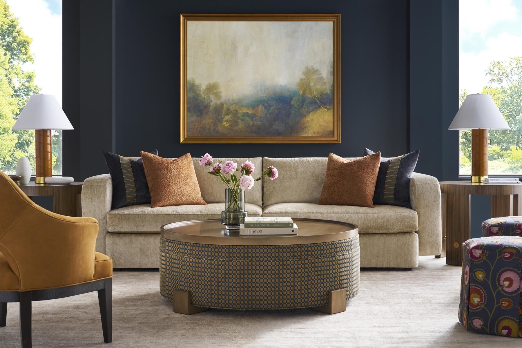 Navy-walled living room with a tufted sofa, round cocktail ottoman and landscape painting