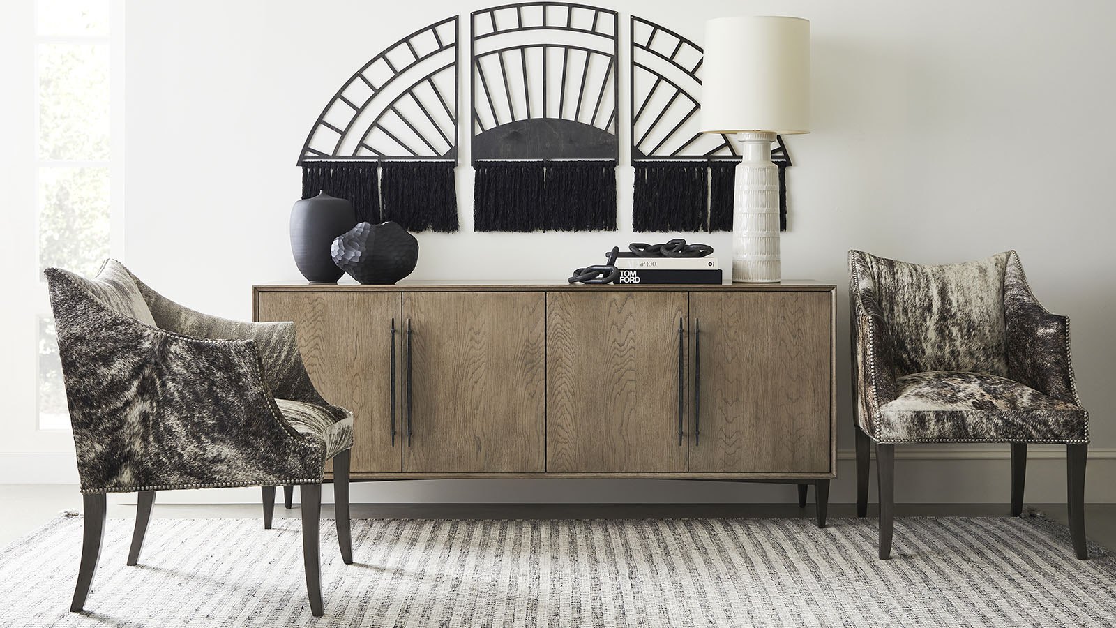 Oak four-door credenza with black pulls between a pair of hide-upholstered chairs, under a black fan wall sculpture